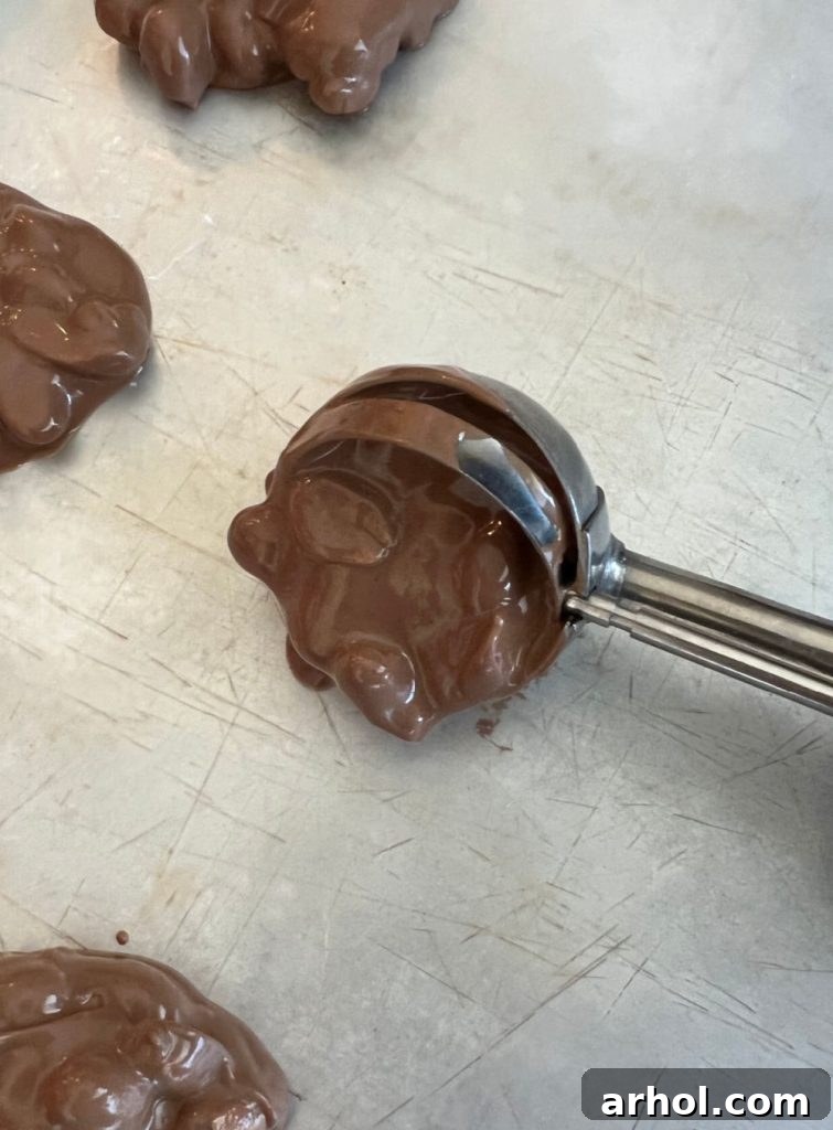 Using a small cookie scoop to place melted chocolate peanut mixture onto a wax paper lined cookie sheet.
