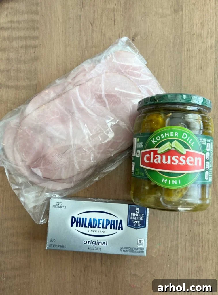 Close-up of thin slices of deli ham, soft cream cheese, and crisp dill pickles laid out, ready for making ham pickle roll ups.
