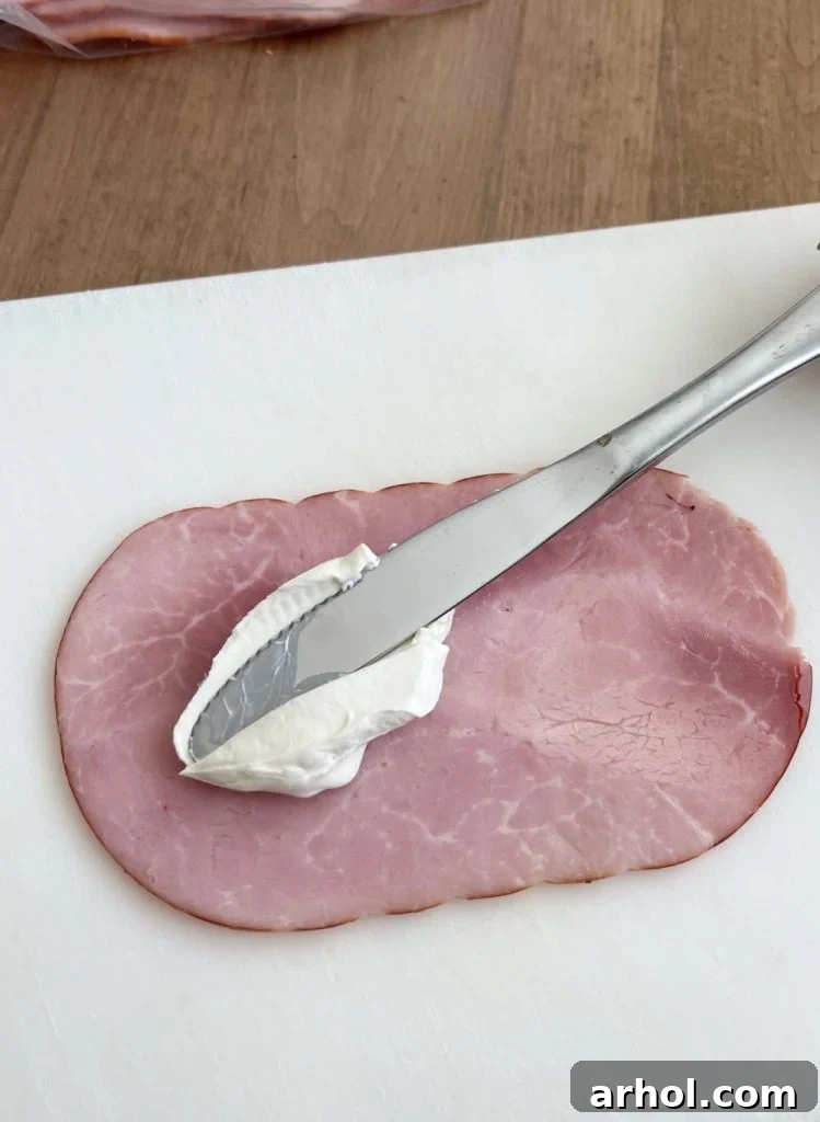 A close-up shot showing cream cheese being spread smoothly onto a slice of deli ham.