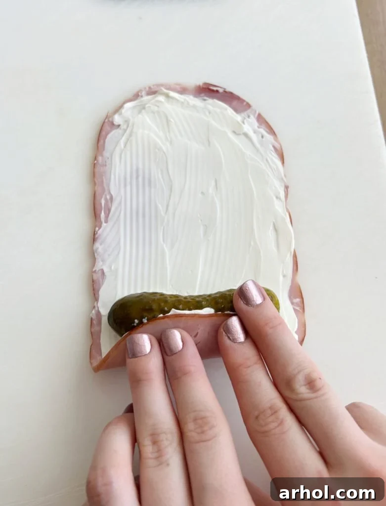 Hands carefully rolling a slice of deli ham with cream cheese around a dill pickle.