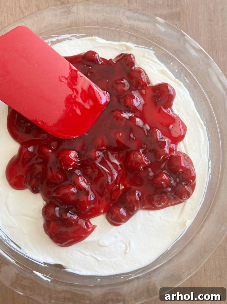 Luscious Cherry Cheesecake Dip 6 Spreading bright red cherry pie filling evenly over the creamy white cheesecake dip in a dish.