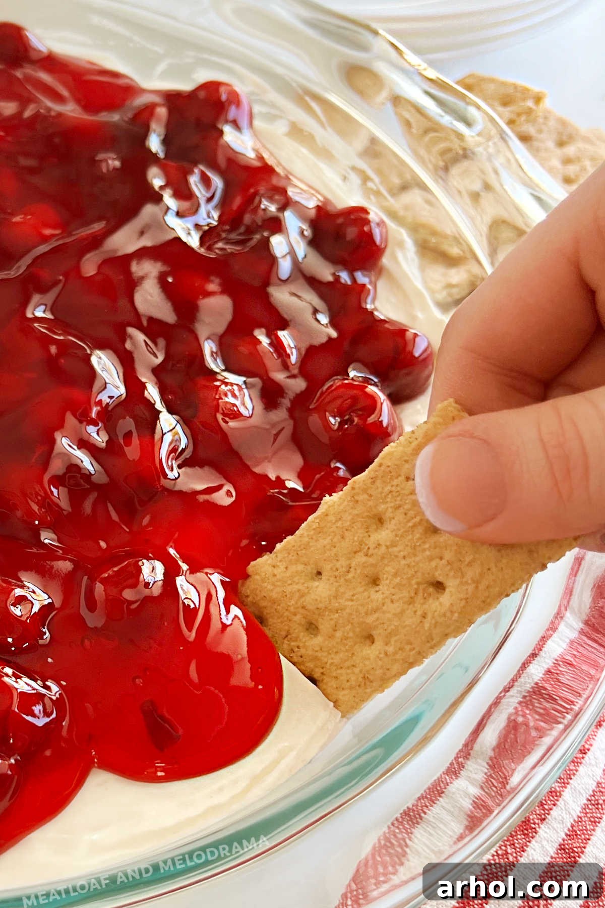 Luscious Cherry Cheesecake Dip 8 A hand dipping a graham cracker into the creamy cherry cheesecake dip, showing the layers of dip and pie filling.