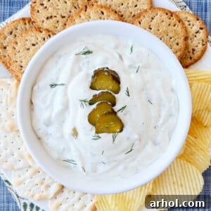 Dill pickle dip with dill pickle slices and fresh dill in a bowl, surrounded by potato chips and crackers