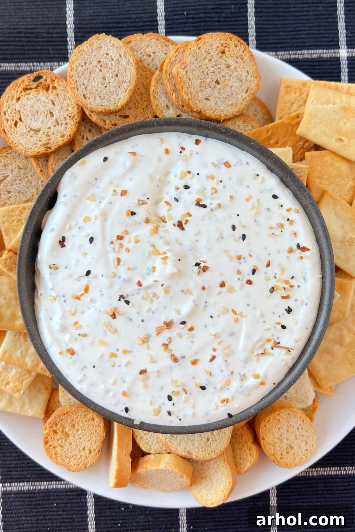 A bowl of creamy Everything Bagel Dip surrounded by an assortment of dippers, including bagel chips, carrots, and celery on a festive platter.