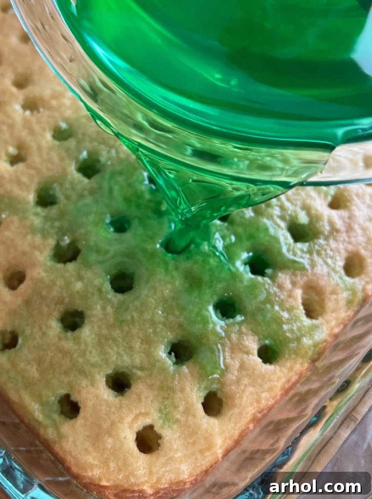 Pouring the bright green lime Jell-O mixture evenly over the poked cake, ensuring all holes are filled.