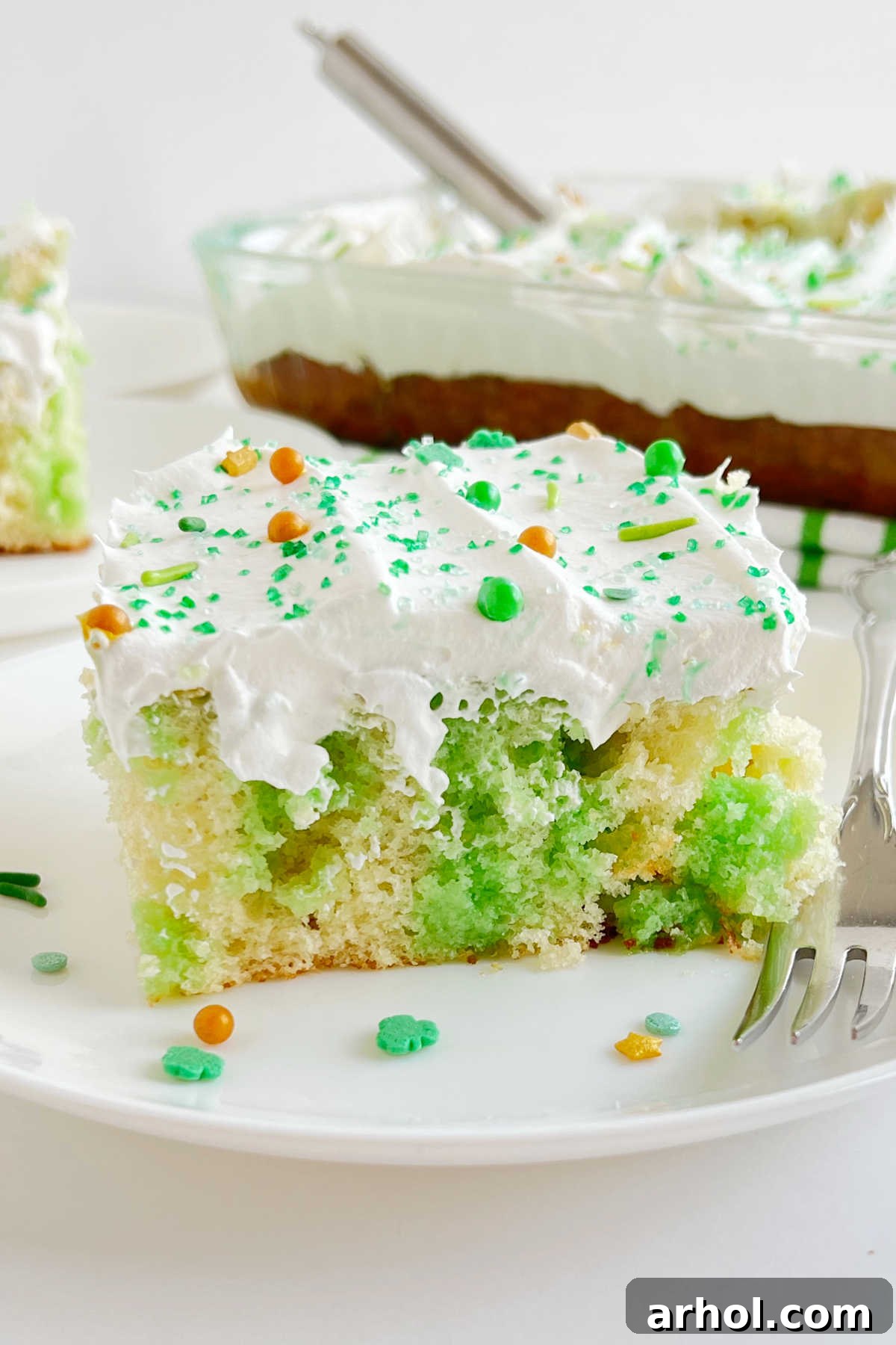 A perfectly sliced piece of Lime Jello Poke Cake, showing the vibrant green Jell-O soaked into the fluffy white cake, topped with creamy Cool Whip and sprinkles, served on a white plate.