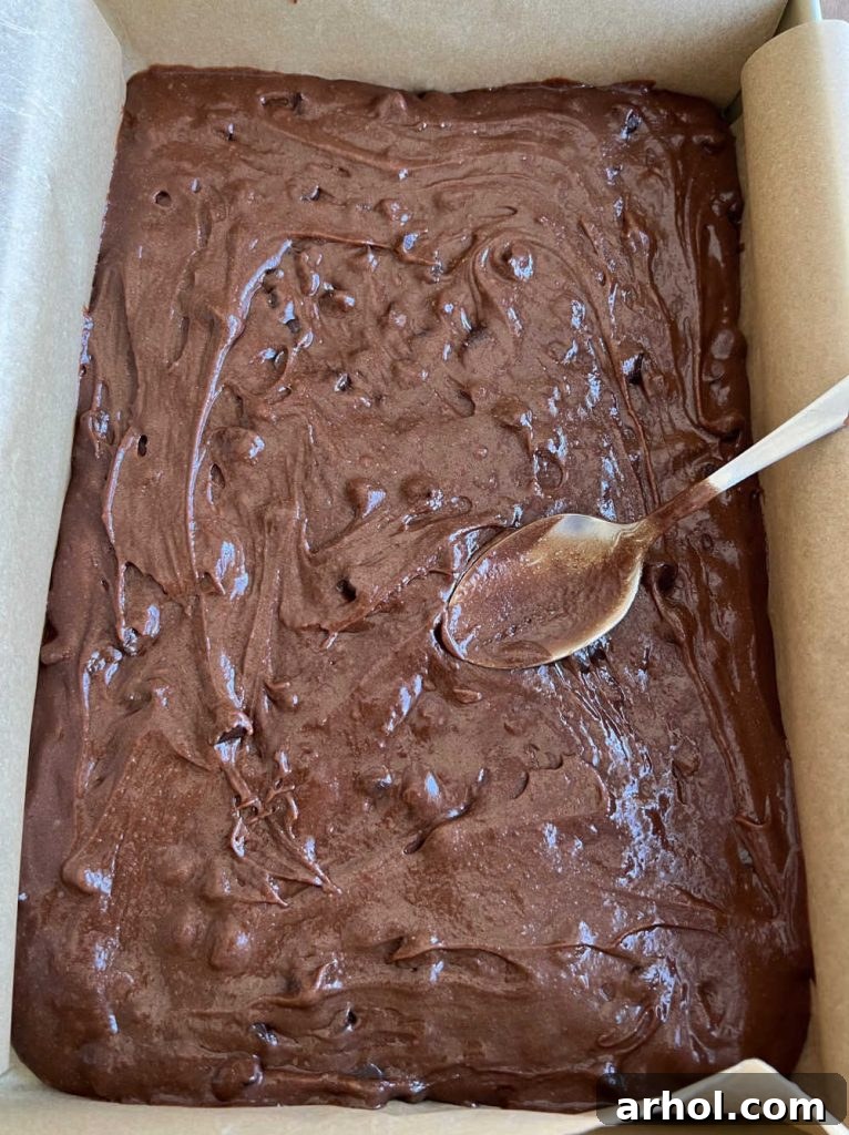 Mini Egg Magic Brownies 5 Thick brownie batter being spread evenly into a parchment paper-lined 9x13 inch baking dish before baking.