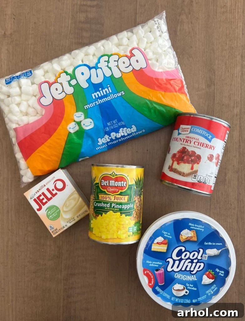Individual ingredients for Cherry Fluff: a can of cherry pie filling, a tub of Cool Whip, a can of crushed pineapple, a box of vanilla pudding mix, and a bag of mini marshmallows, laid out on a white surface.
