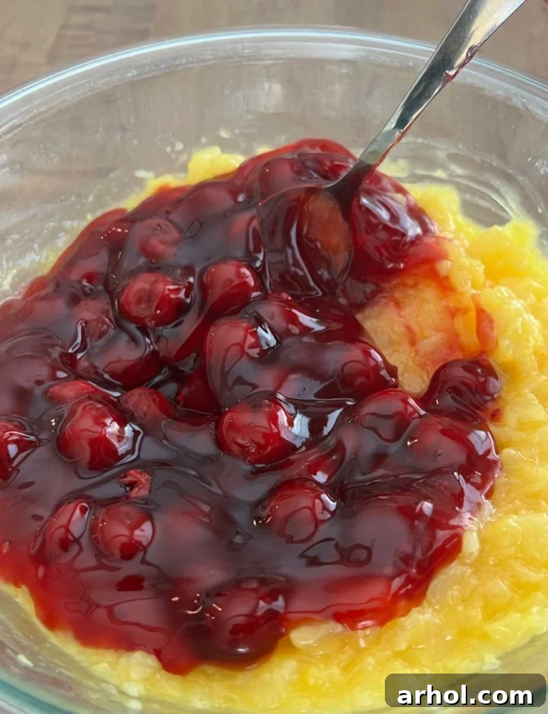 Cherry pie filling being stirred into the pineapple and vanilla pudding mixture in a large mixing bowl, creating a vibrant pink base for the fluff.