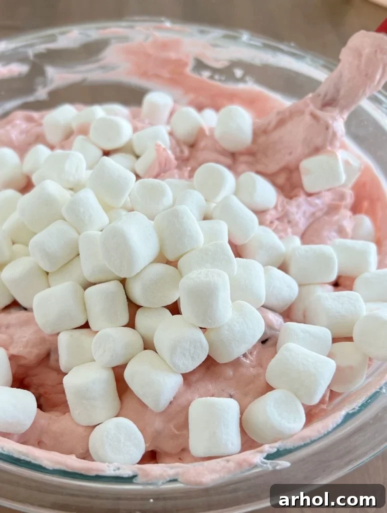 The final step: gently folding thawed whipped topping and mini marshmallows into the pink cherry and pineapple mixture in a bowl.