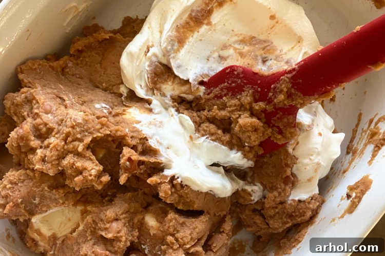 mix refried beans with cream cheese and sour cream in white baking dish for bean dip preparation