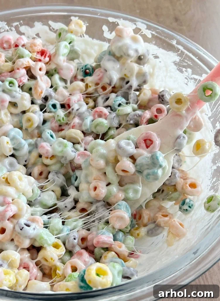 A rubber spatula expertly stirring colorful Froot Loops cereal into a bowl of gooey, melted marshmallow mixture, ensuring every piece is coated.