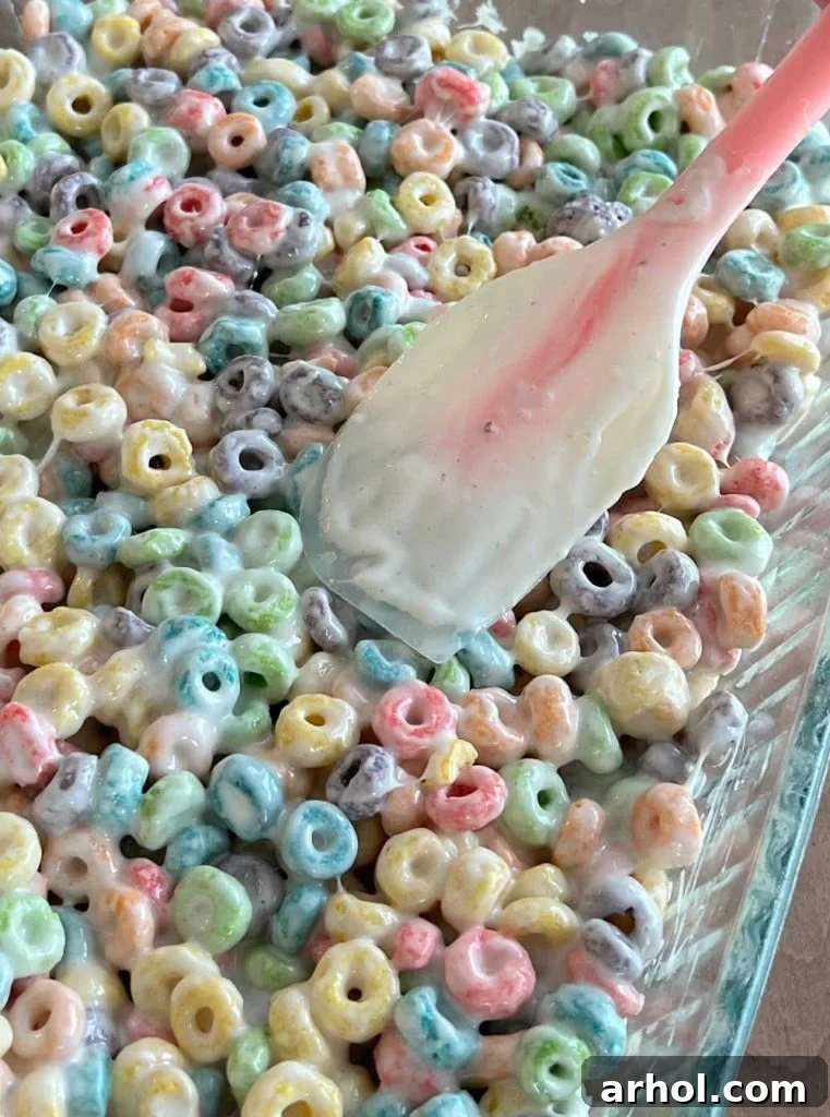 The vibrant Froot Loop cereal mixture being gently patted and spread into a glass baking dish with a spatula, creating an even layer for setting.