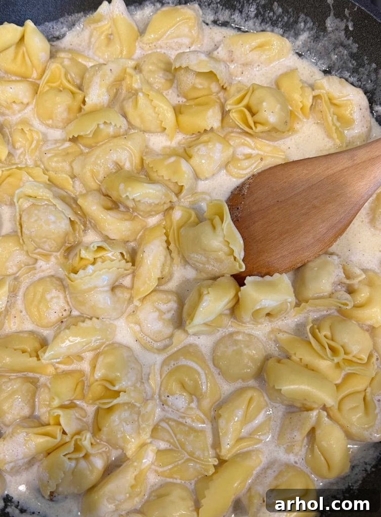 Fresh cheese tortellini being stirred into the creamy Parmesan sauce in a large skillet with a wooden spoon.