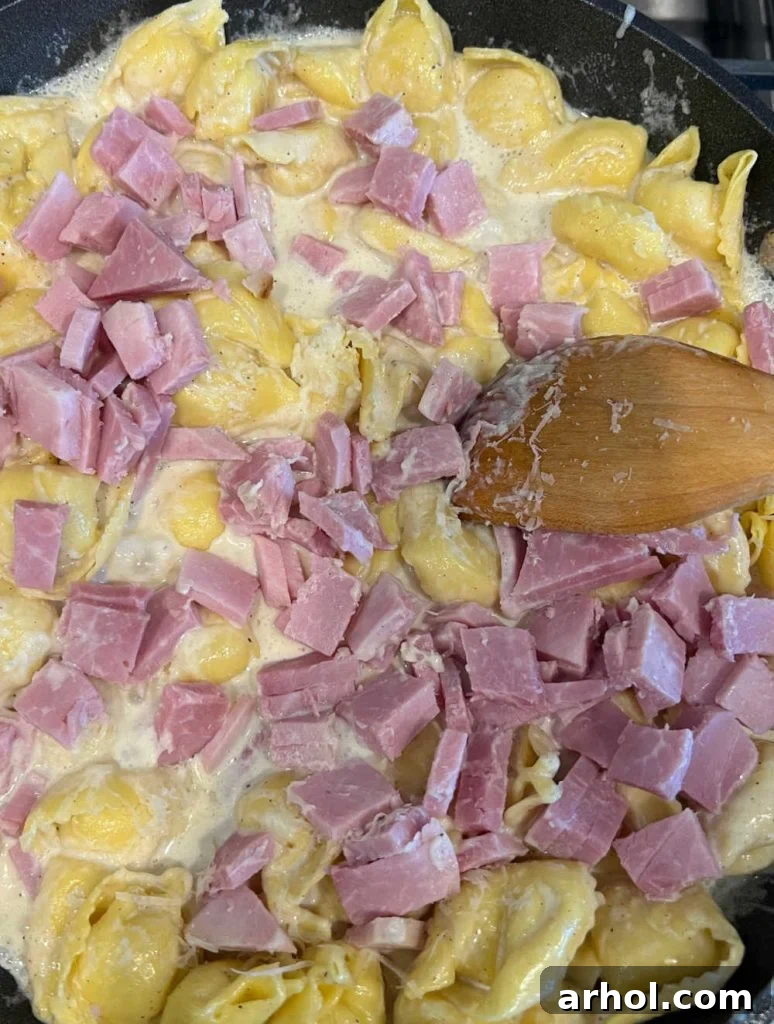 Diced cooked ham being stirred into a skillet with cooked tortellini and creamy sauce.