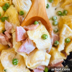 Creamy tortellini with ham and peas in a Parmesan cheese sauce on a wooden spoon