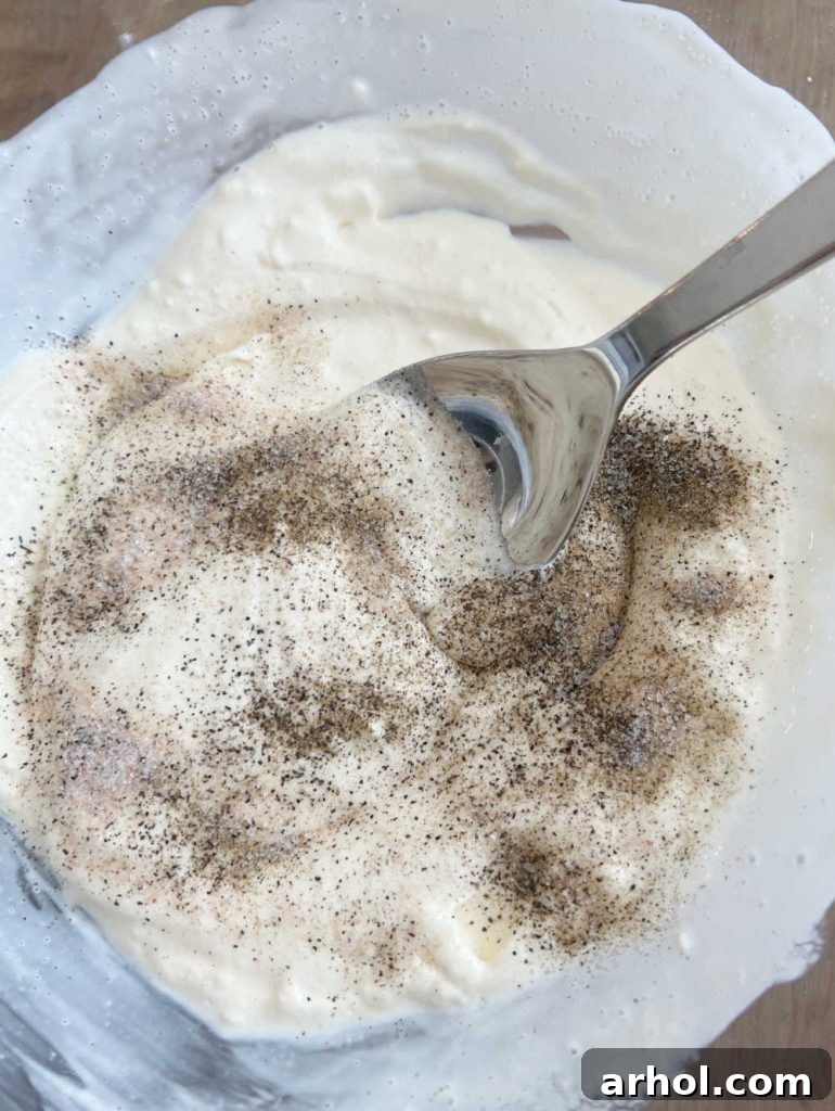 The process of mixing the creamy dressing for Hawaiian macaroni salad: mayonnaise, milk, salt, and pepper are vigorously stirred together in a large white bowl.