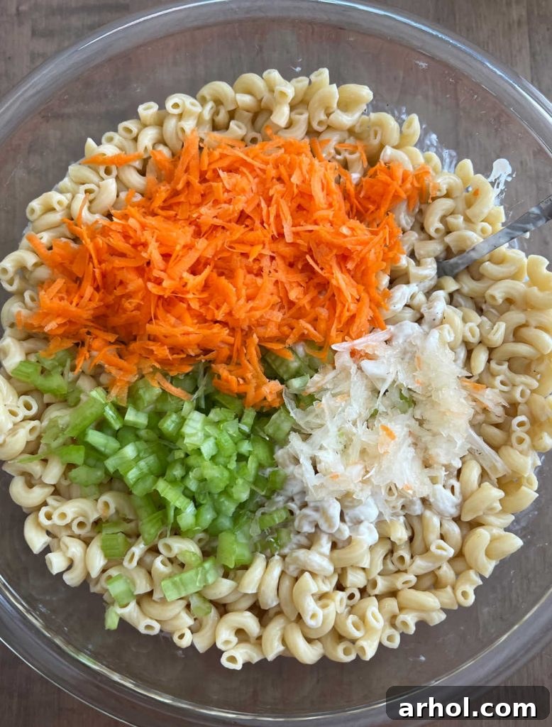 A large mixing bowl filled with cooked elbow macaroni, shredded carrots, finely diced celery, and grated onions, all ready to be tossed with the creamy dressing to form Hawaiian mac salad.