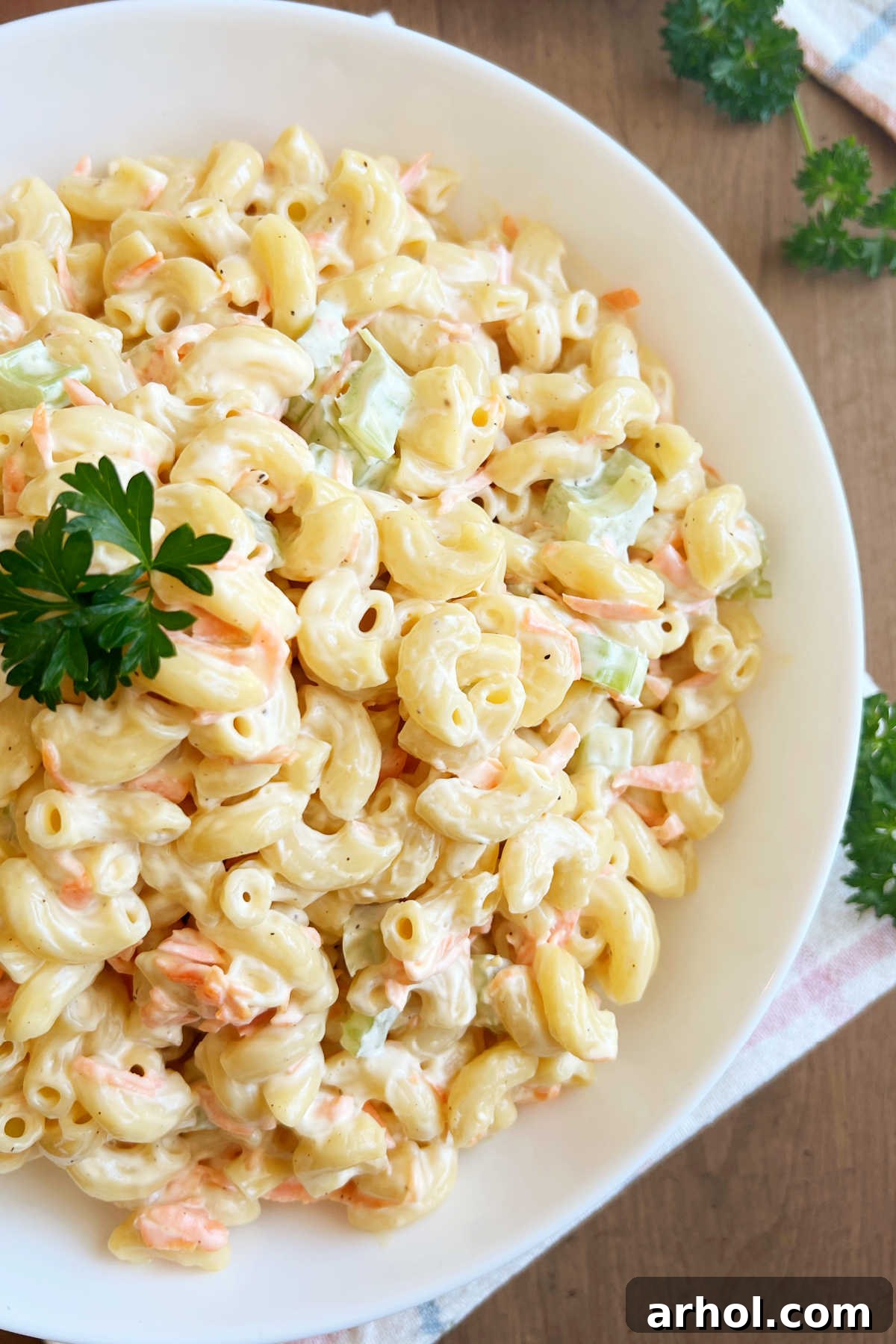 A beautifully presented bowl of creamy Hawaiian mac salad, garnished with fresh green parsley, resting on a clean white surface with a serving spoon nearby.