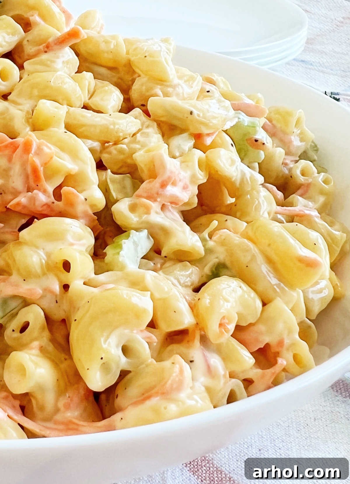 A close-up side view of Hawaiian macaroni salad, showcasing its creamy texture, vibrant grated carrots, and diced celery in a white bowl on a wooden table.