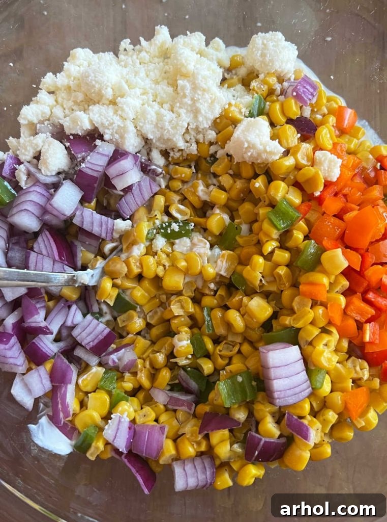 All the vibrant ingredients for Mexican street corn salad, including the charred corn mixture, diced peppers, onions, cilantro, and Cotija cheese, being mixed thoroughly with the creamy dressing in a large bowl.
