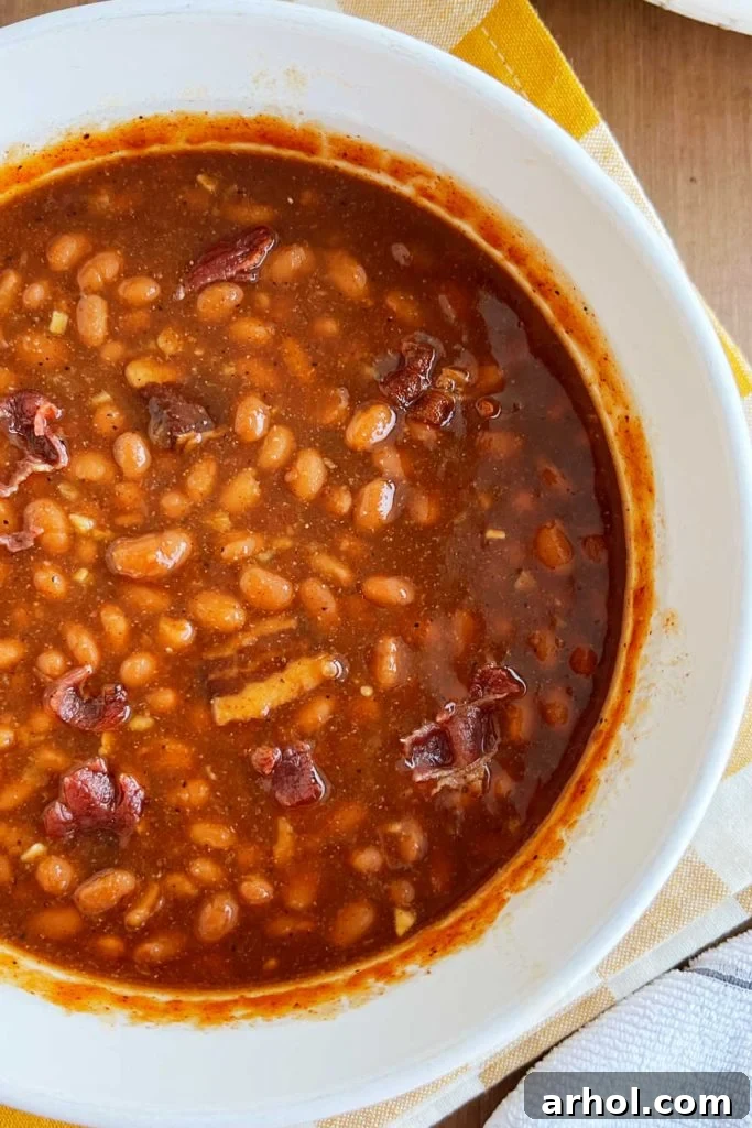 A close-up shot of rich, dark brown sugar baked beans in a white casserole dish, garnished with crispy bacon, showcasing a delectable texture.