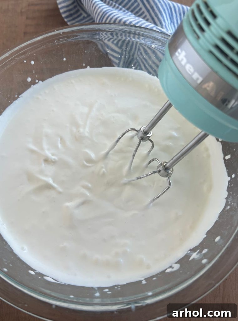 No-Churn Chocolate Peanut Butter Bliss 4 Heavy whipping cream being whipped in a large metal bowl with an electric mixer until stiff peaks form.