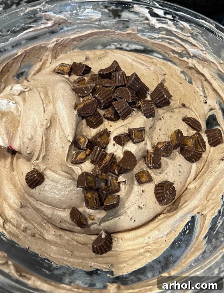 No-Churn Chocolate Peanut Butter Bliss 6 Chopped Reese's candies being gently folded into the creamy whipped cream and peanut butter mixture with a spatula.