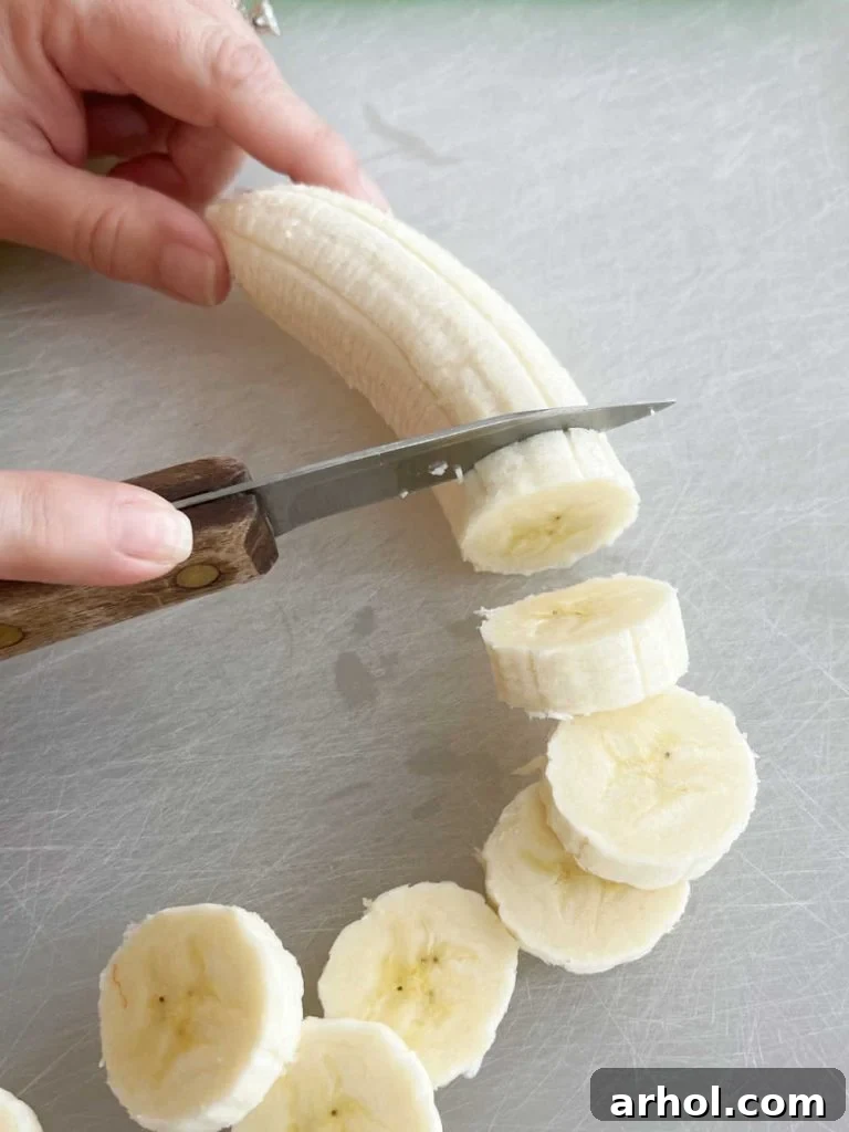 A hand using a knife to cut a ripe banana into uniform slices on a cutting board, preparing for dipping.