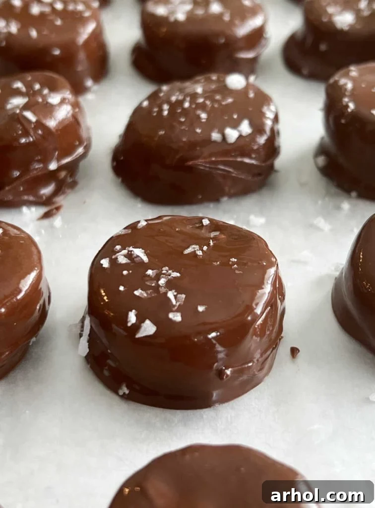 A close-up shot of flaked sea salt being sprinkled over freshly chocolate-covered banana slices arranged on parchment paper.