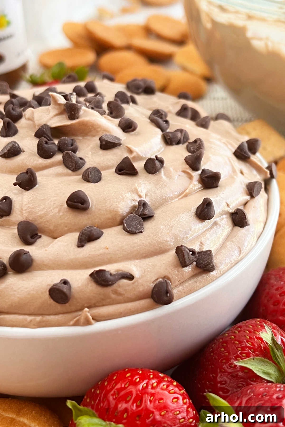 A beautiful bowl of creamy Nutella cheesecake dip, generously garnished with mini chocolate chips, surrounded by an assortment of fresh strawberries and vanilla wafer cookies, ready for dipping.