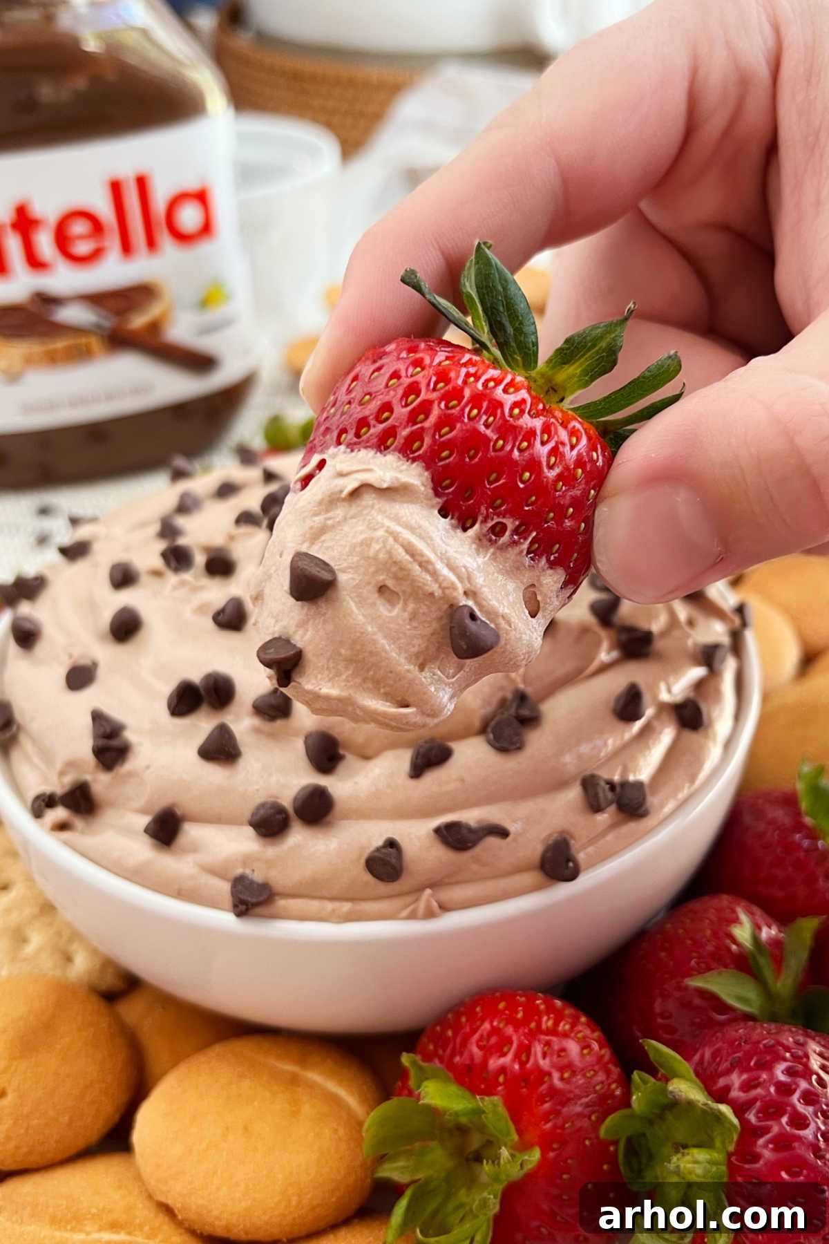 A vibrant, fresh strawberry being perfectly dipped into a bowl of creamy Nutella cheesecake dip, which is highlighted with a sprinkle of mini chocolate chips, creating an enticing visual.