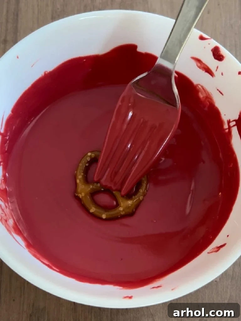 Dipping a mini pretzel into melted red candy coating, showing the gentle tapping technique to remove excess.