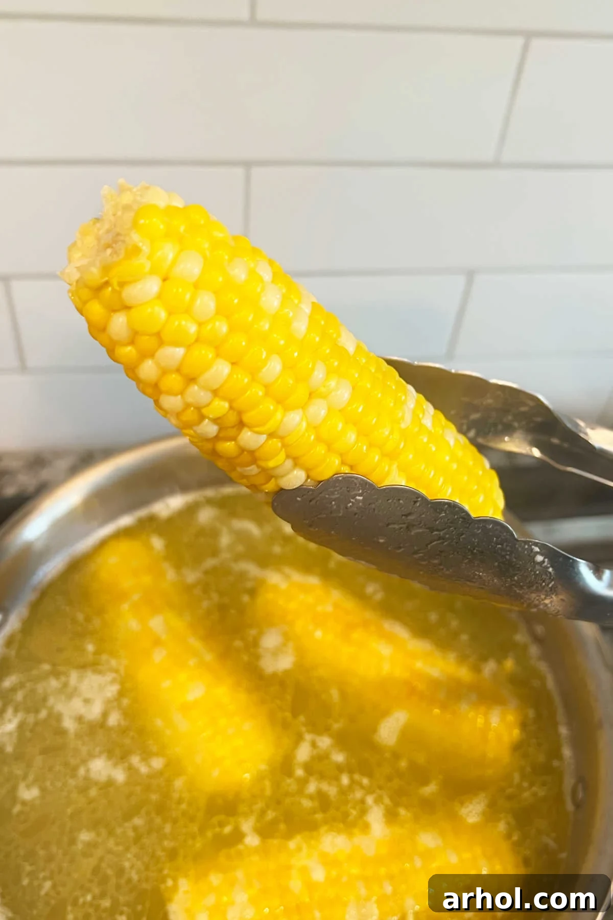 Ear of perfectly boiled corn on the cob held by kitchen tongs over a pot of boiling water with melting butter, ready to be served.