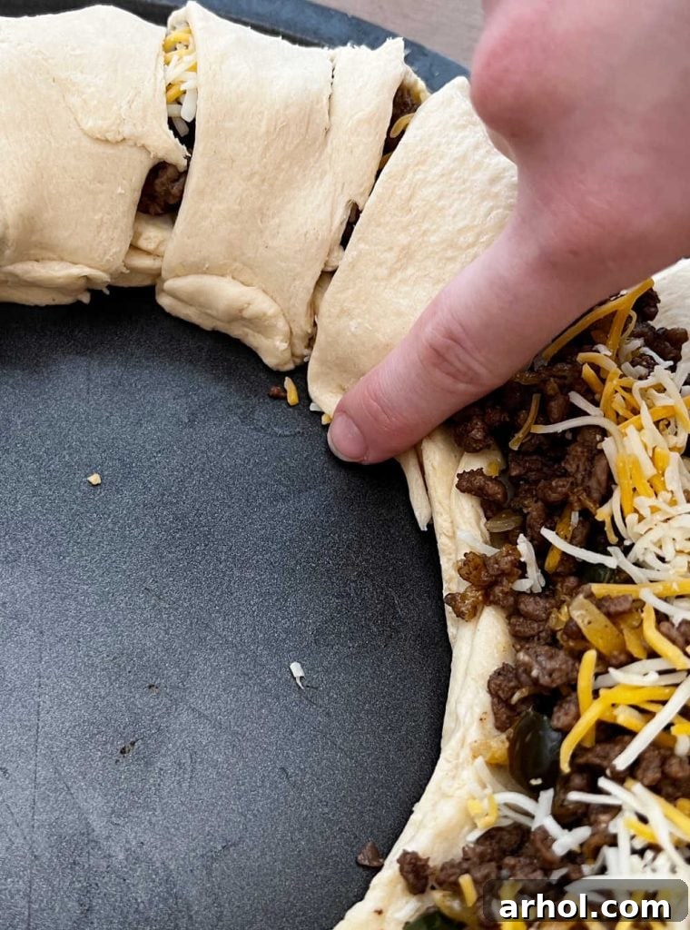Hand folding the tip of a crescent roll triangle over the taco meat filling, pressing it to seal the ring.