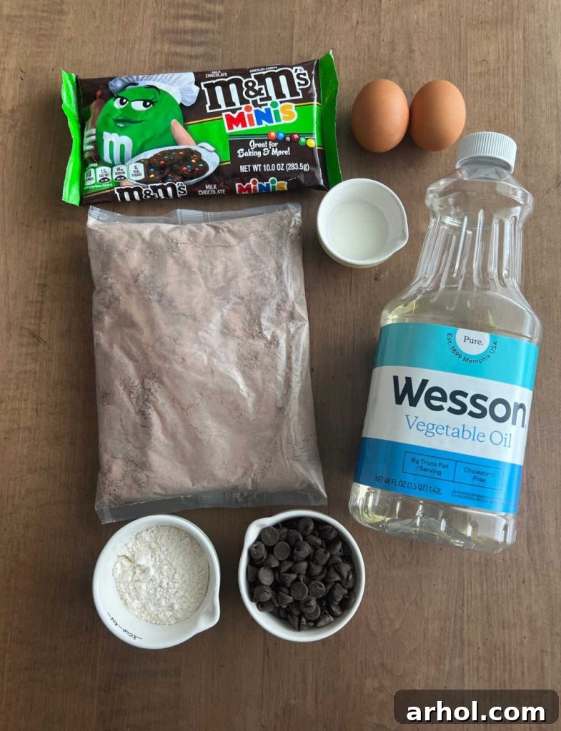 A vibrant flat lay showcasing all the essential ingredients needed for brownie mix cookies: a box of brownie mix, colorful M&M minis, fresh eggs, milk, vegetable oil, all-purpose flour, and rich chocolate chips.