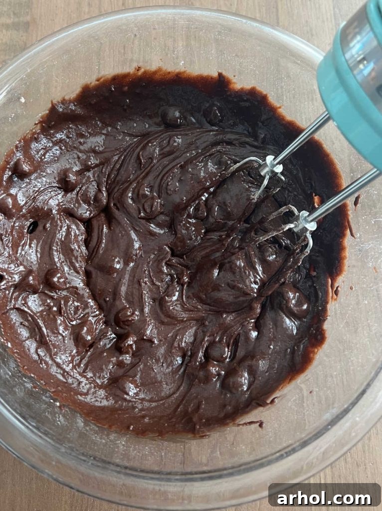 Rich brownie batter in a mixing bowl being thoroughly combined with an electric mixer, illustrating the smooth and thick consistency before adding mix-ins.