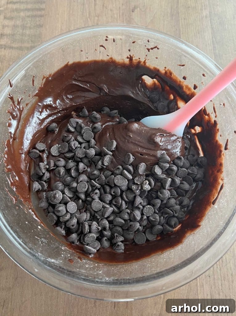 A spatula gently folds generous semi-sweet chocolate chips into the thick, dark chocolate batter in a mixing bowl, showcasing the final stage of mixing before baking.