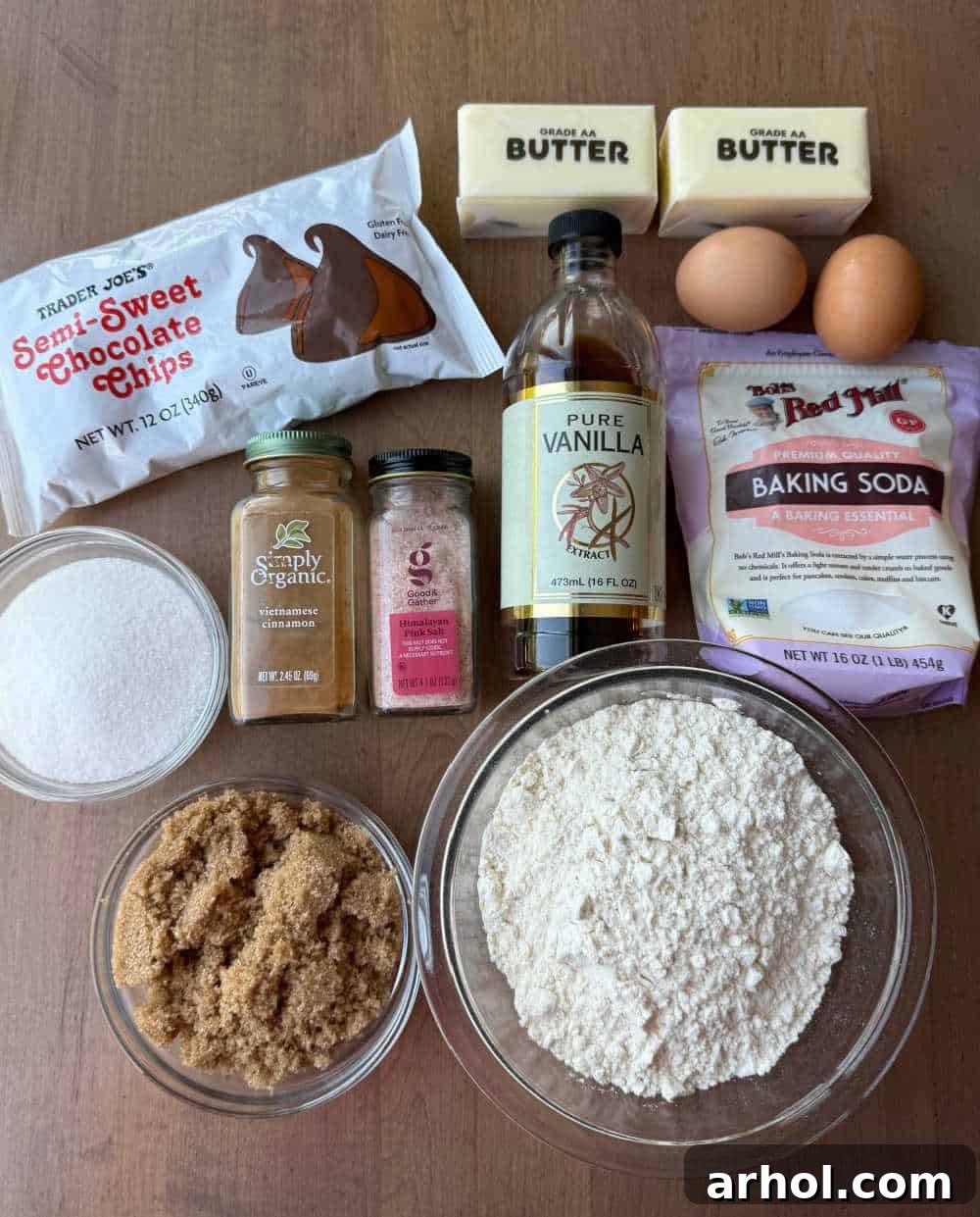 Ingredients for chocolate chip cookies laid out on a wooden table, including flour, sugars, butter, eggs, and chocolate chips.