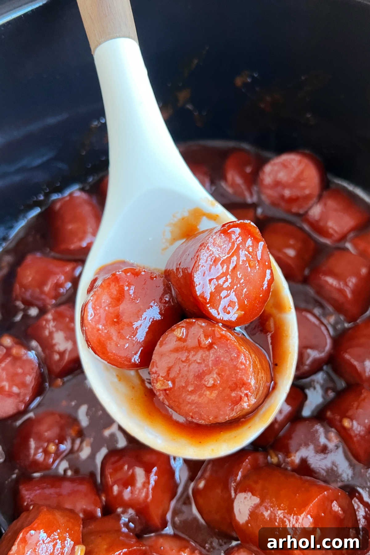 Serving spoon lifting a piece of slow cooker kielbasa bite with BBQ sauce from a crock pot, illustrating its deliciousness and glossy glaze.
