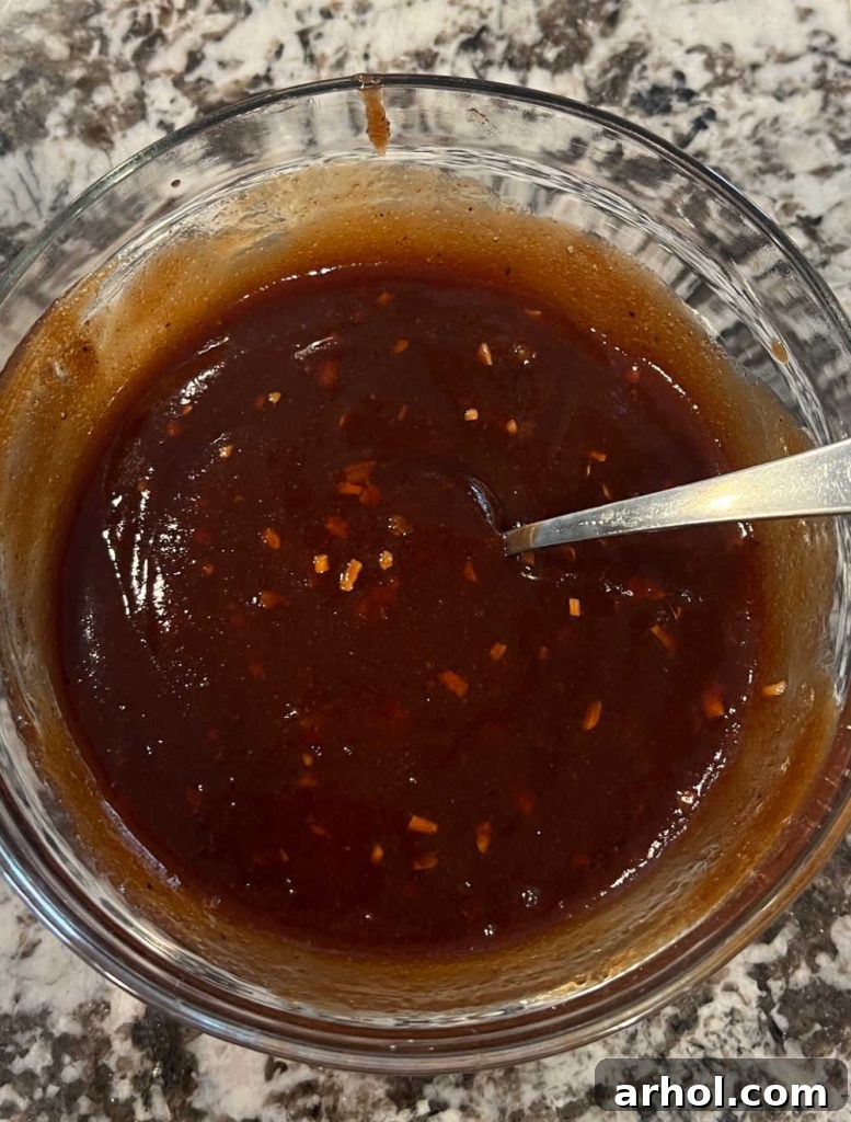 Close-up of BBQ sauce, brown sugar, and other spices being mixed in a small glass bowl with a spoon, showing the sauce's texture.