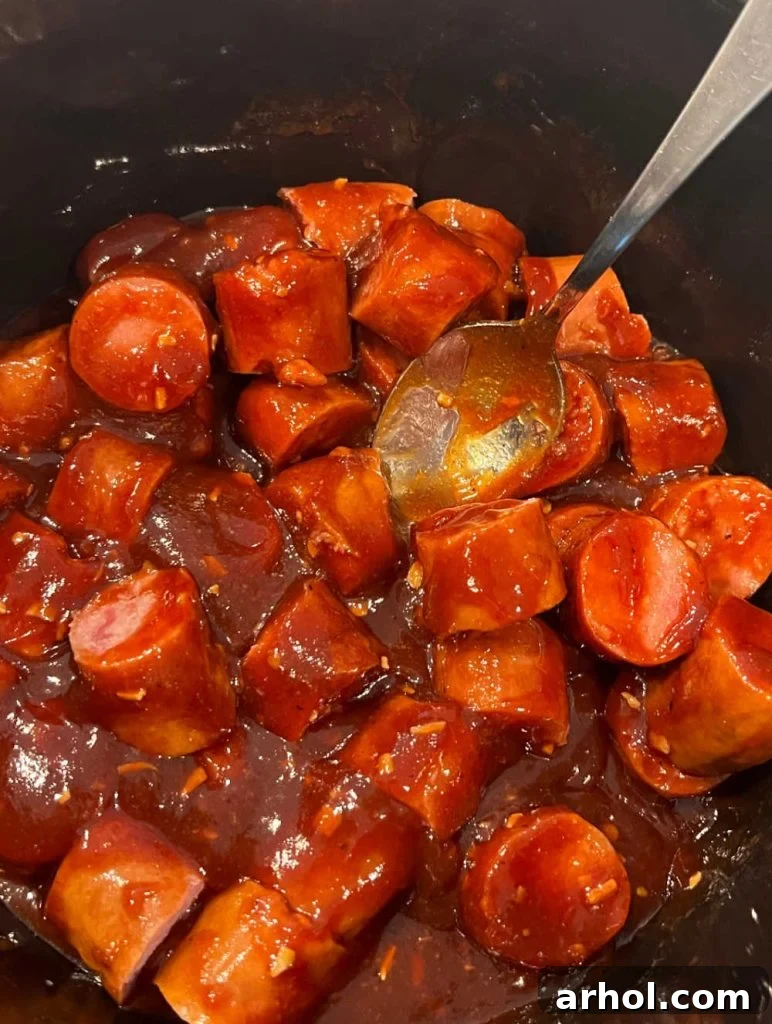 Kielbasa slices and rich BBQ sauce being stirred together in a slow cooker with a serving spoon, showcasing the even coating before cooking begins.