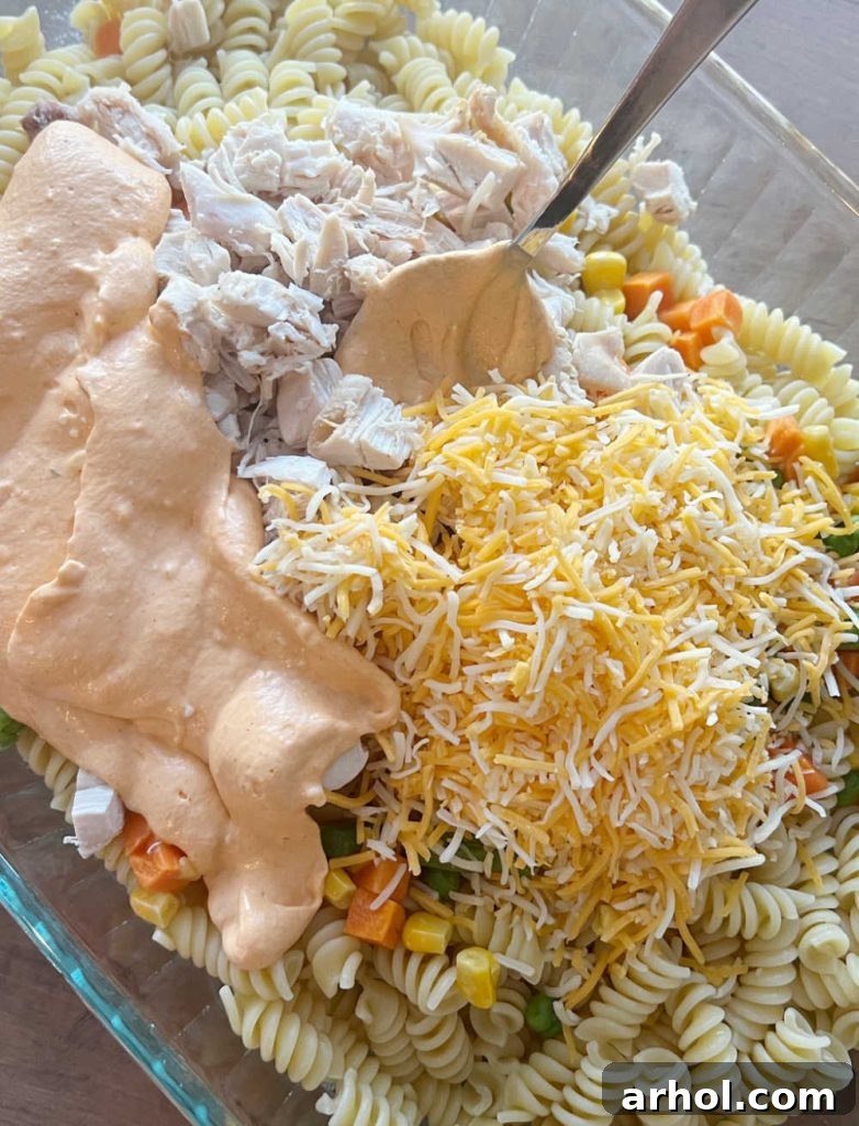 Assembling the Buffalo Chicken Casserole in a baking dish, showing mixed pasta, chicken, vegetables, and sauce before topping with cheese.