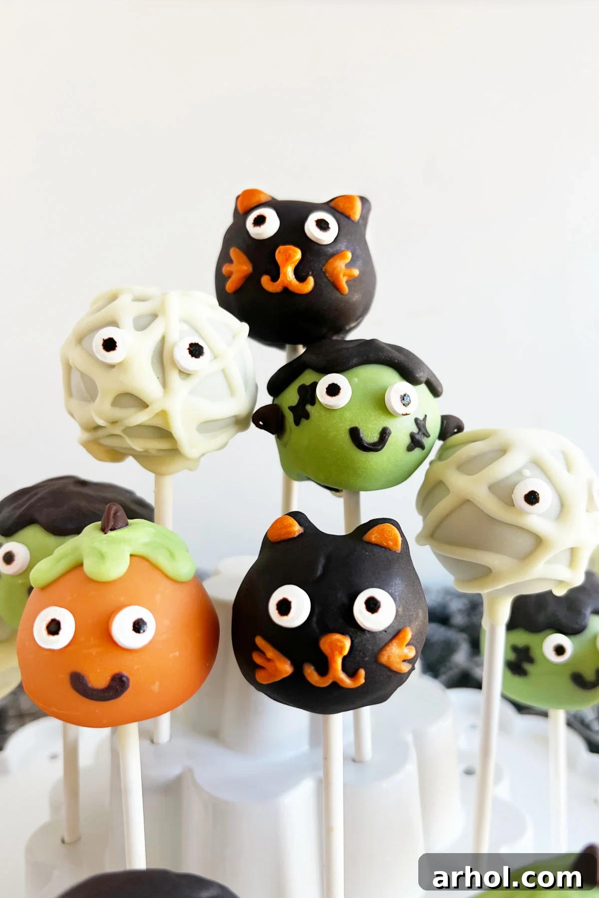 Halloween cake pops decorated as a friendly black cat, a classic mummy, a grinning pumpkin, and a quirky Frankenstein monster.
