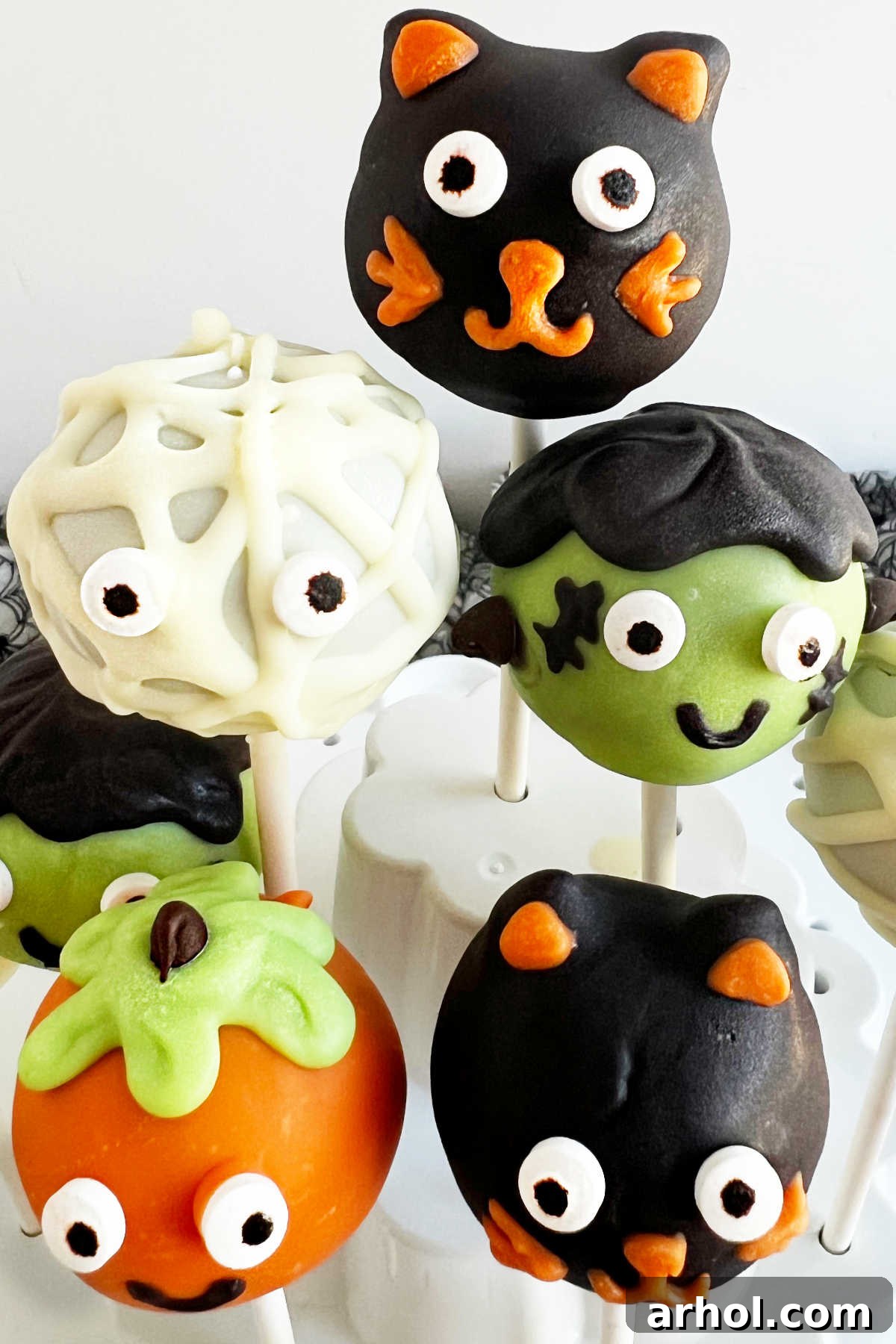 A close-up view of an assortment of Halloween cake pops, including a mummy, pumpkin, and Frankenstein, displayed artfully in a cake pop holder.