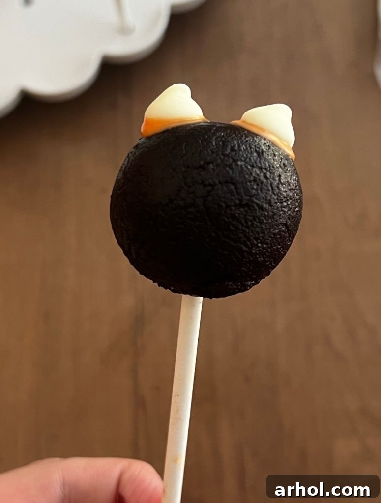 Instructions for creating black cat cake pops, showing the attachment of chocolate chip ears and then dipping in black candy melt.