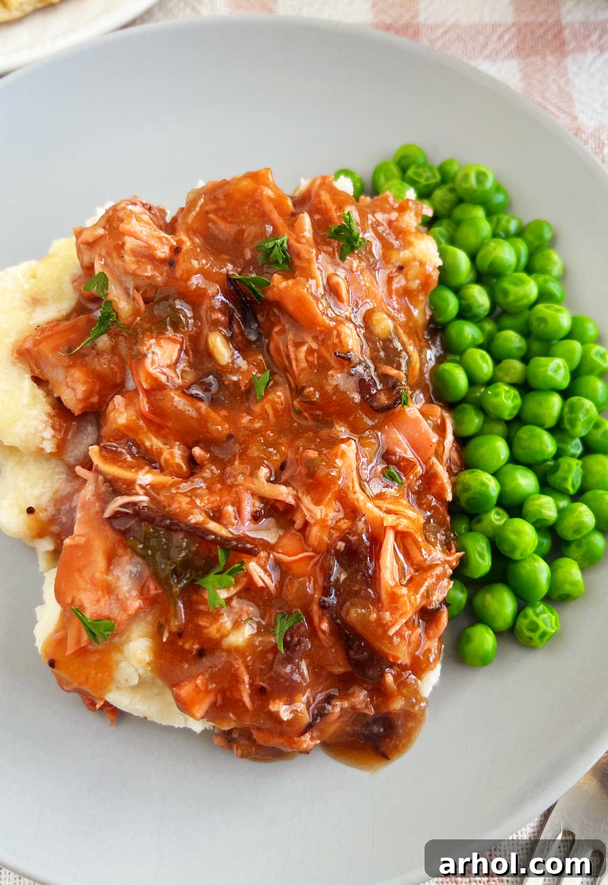 Shredded slow cooker cranberry chicken served alongside creamy mashed potatoes and vibrant green peas on a white plate.
