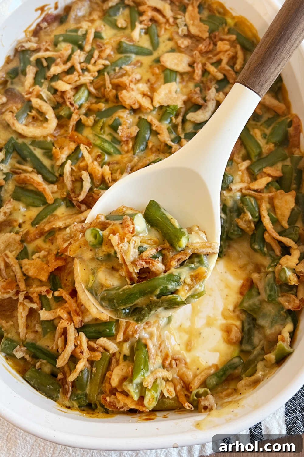 A generous serving of Campbell's Green Bean Casserole, adorned with golden-brown French's fried onions, presented on a white serving spoon. Steam gently rises, hinting at its warmth and creamy texture.
