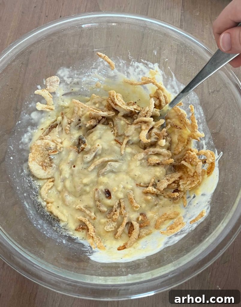 Green beans and half a cup of crispy fried onions are stirred into the creamy soup mixture in a large mixing bowl, ensuring all ingredients are well combined before baking.