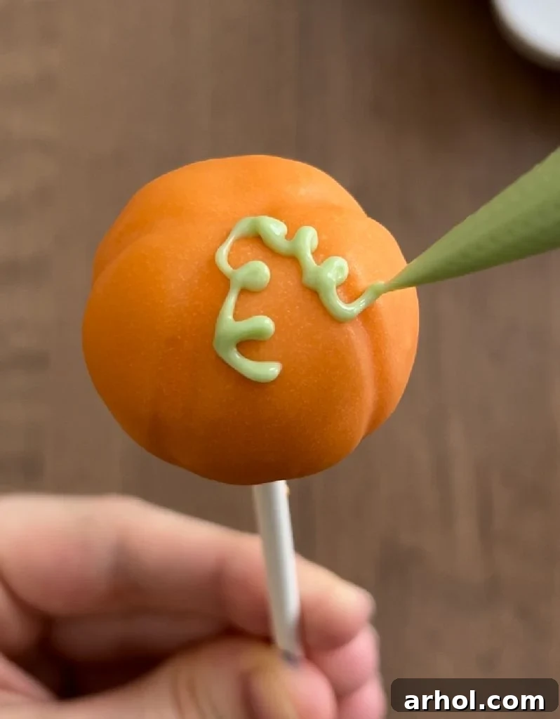 A pumpkin cake pop being decorated with a green piping bag to create a delicate vine design.